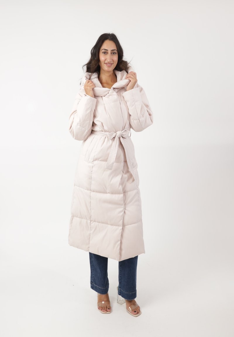 Abrigo acolchado en rosa claro, con un cuello alto y cinturón, que presenta secciones acolchadas y una textura suave. Combinado con vaqueros y tacones de bloque transparentes.