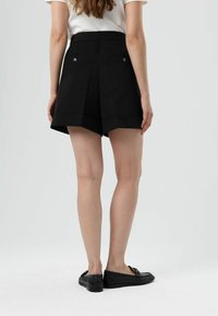 Shorts noirs sur mesure à taille haute, avec des plis et deux poches arrière, portés avec des chaussures noires sans lacets et un haut blanc.