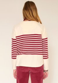 Femme de dos portant un haut à manches longues crème avec des rayures horizontales bordeaux et un pantalon en velours côtelé bordeaux, sur fond uni.