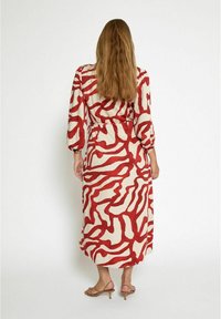 Langsleeve jurk in crème met gedurfde rode abstracte patronen, ceintuurovergang, en een vloeiende silhouette. Glad stof met een lichte glans.