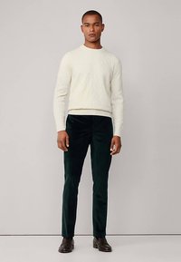 Kermanvärinen palmikkoneulepaita yhdistetty tummanvihreisiin samettihousuihin. Malli seisoo suorana, jalassa ruskeat nahkakengät, korostaen tyylikästä istuvuutta.