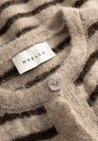 Îmbrăcăminte tricotată cu dungi bej deschis și maro, cu decolteu rotund, nasture și etichetă albă cu inscripția „NOELLA” cusută în interiorul gâtului.