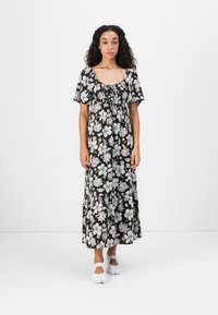 Sort blomster maxi kjole med hvide blomsterm�ønstre, korte ærmer og en afslappet pasform. Båret med hvide sko, stående på en ensfarvet baggrund.