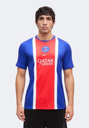 Homme portant un maillot de football bleu et rouge du Paris Saint-Germain avec le logo Qatar Airways, debout face à l'avant sur un fond uni.