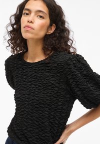 Melna teksturēta blūze ar sakulšanas dizainu, apaļu kakla izgriezumu un īsām pūkainām piedurknēm. Audumam ir sarautas izskats.