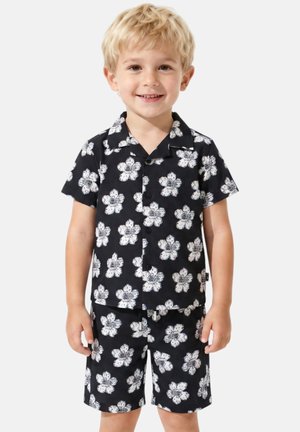 Tout-petit blond souriant, portant un t-shirt noir à manches courtes et un short à imprimé floral blanc, debout sur un fond clair uni.