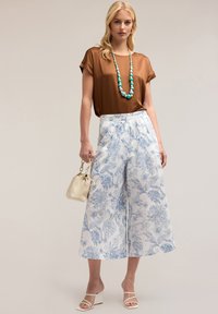 Brun short-sleeve top, valkoiset leveälahkeiset housut, joissa sininen kukkakuosi, valkoiset hihnalliset korkokengät ja pieni beige laukku. Helmikaulakoru mukana.