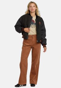 Femeie îmbrăcată într-o jachetă bomber neagră și gri, peste un tricou grafic bej, pantaloni din catifea lățiti maro și adidași negri, stând cu spatele la un fundal alb.