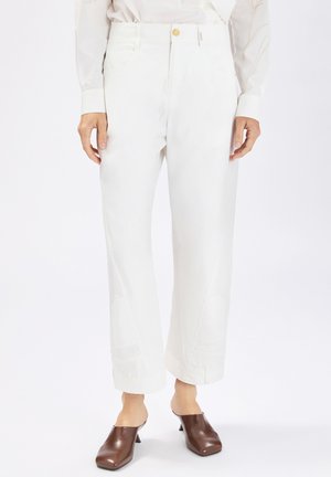 Pantaloni bianchi a gamba dritta con tasche frontali e chiusura con bottone, indossati con mule con tacco marroni e una camicia a maniche lunghe di colore chiaro.