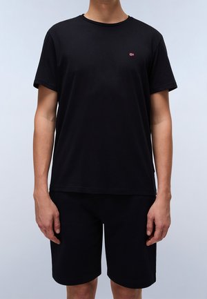 Zwarte katoenen T-shirt met korte mouwen en een ronde hals, met een klein rood en wit logo op de linkerborst. Gecombineerd met zwarte shorts.