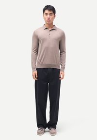 Mann steht aufrecht, trägt ein beigefarbenes Langarm-Poloshirt, lockere schwarze Hosen und hellgraue Schnürschuhe vor einem schlichten Hintergrund.