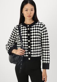 Svart och vit stickad cardigan med ett hjärtformat mönster, rund hals, knappstängning och ribbade ärmslut. Modellen håller en svart väska.