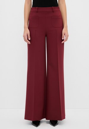 Pantalons larges en tissu bourgogne, avec deux poches, une taille ajustée et une texture lisse. Portés avec un haut noir ajusté.