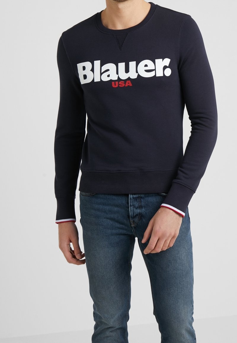Homme portant un sweat-shirt bleu marine avec l'inscription "Blauer USA" en grandes lettres blanches et rouges, associé à un jean bleu, debout devant un fond uni.
