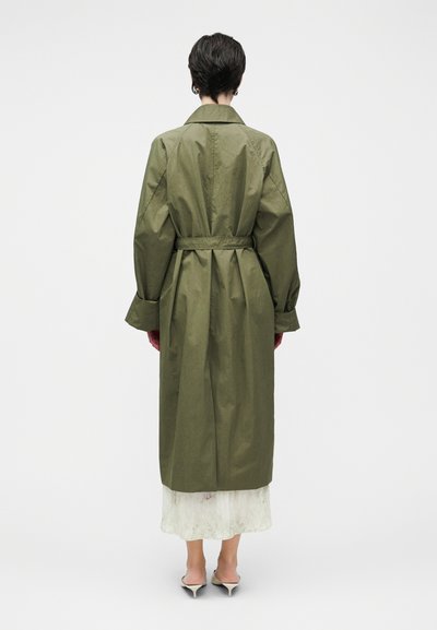 Λαδί μακρύ παλτό με ζώνη στη μέση, φαρδιά μανίκια και λεία υφή, φορεμένο πάνω από ένα ελαφρύ, στρωματοποιημένο ένδυμα.