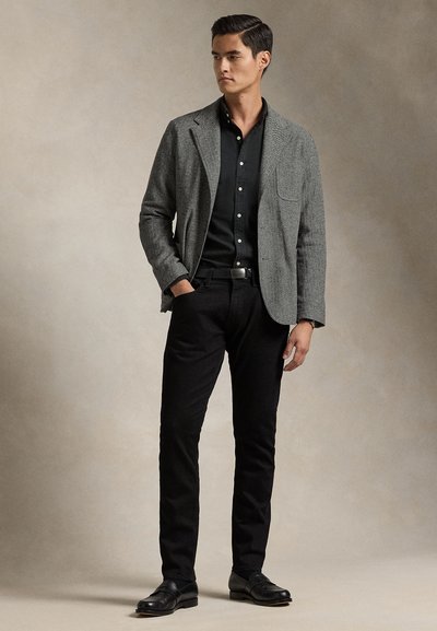 Moški stoji z rokami v žepih, oblečen v siv blazer, črno srajco na gumbe, črne hlače in črne elegantne čevlje na enobarvnem ozadju.