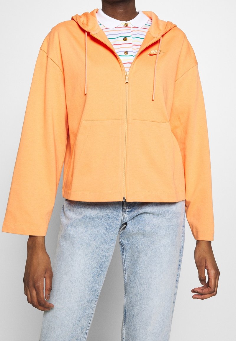 Kangaskaputta, joka on kirkkaan oranssi, pehmeästä kankaasta valmistettu, etutasku ja nyörit, puettuna raidallisen polo-paidan päälle.