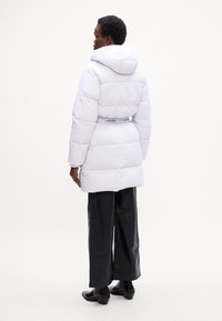 Biela puff jacket s kapucňou, prešívaný dizajn, sťahovaný pás a kontrastný šedý branding detail na opasku. Nosené s čiernymi nohavicami.