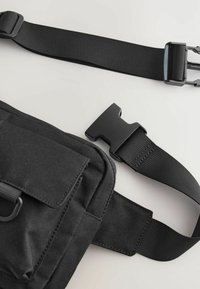 Black messenger bag with a textured fabric, adjustable strap, and black plastic buckle closure. Features a front flap pocket and metal D-ring.