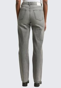 Person i högmidjade, raka grå jeans och svarta klumpiga skor, stående med ryggen mot kameran mot en enfärgad bakgrund.