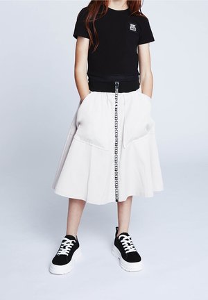 T-shirt noir court avec logo, associé à une jupe gris clair dotée de poches, d'un détail de zip rayé noir et de baskets noir et blanc.