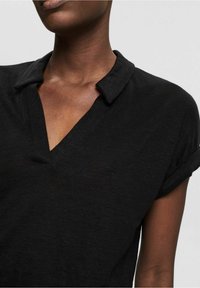 Gros plan d'une personne portant une chemise noire à manches courtes avec un col plié et un décolleté en V.