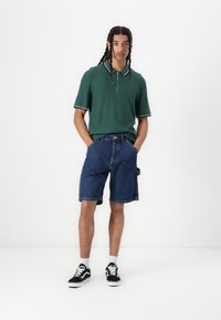 Polo shirt verde con texture e colletto a zip, abbinato a pantaloni corti in denim blu fino al ginocchio e sneakers nere con suola bianca.
