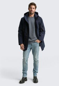 Parka azul marino con capucha, que cuenta con bolsillos con cremallera, combinada con un suéter gris y jeans de un azul claro, finalizada con botas negras.