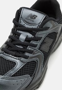 Chaussure de course noire avec une tige en maille et des accents synthétiques. Elle possède une languette rembourrée, des lacets et un logo "N" visible sur le côté.