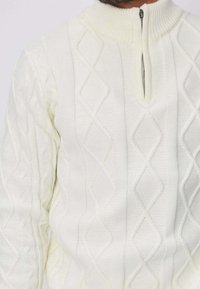 Pull-over en tricot à torsades blanc avec un col montant et une fermeture éclair quart de tour, présentant un motif en losanges et des accents côtelés sur le col.