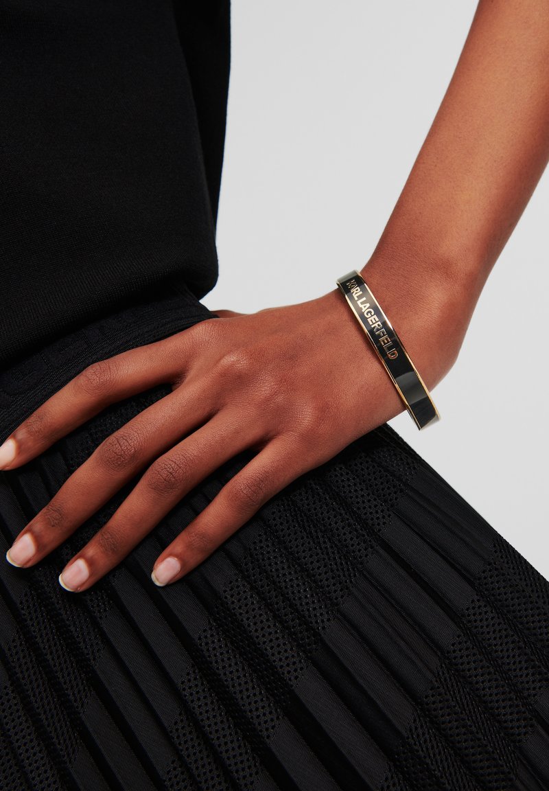 Bracelet doré et noir avec une finition brillante, arborant un logo gravé. Main reposant sur un tissu plissé noir texturé.