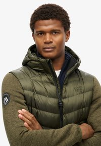 Gilet trapuntato verde oliva con cappuccio, chiusura zip e logo Superdry sul petto e sulla manica. Presenta maniche in maglia verdi.