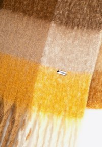 Wełniany szalik w kratę w odcieniach brązu, beżu i musztardowej żółci, z frędzlami na krawędziach i małą metką z napisem „WITTCHEN”.