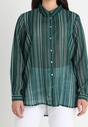 Kobieta ubrana w długą, ciemnozieloną koszulę na guziki z pionowymi białymi paskami, noszącą białe spodnie.