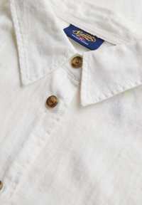 Wit linnen shirt met een puntkraag, twee bruine plastic knopen en een blauw label met de tekst "Superdry". Zachte textuur en casual ontwerp.
