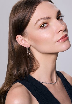 Srebrne kolczyki w kształcie obręczy mają skręcony wzór. Modelka ma długie brązowe włosy i nosi prostą srebrną naszyjnik na tle ciemnej bluzki.