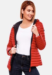 Rode pufferjack met een zwarte binnenkant, met een ritsluiting, een capuchon en een gestikt quiltontwerp; gedragen over een witte shirt en donkere jeans.