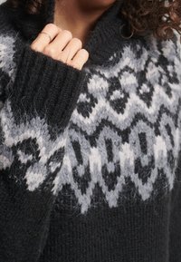 Persoon draagt een zwart-grijs gebreide trui met patroon, hand bij de kraag die een dunne gouden ring aan de vinger toont.