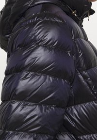 Mørkeblå pufferjakke med en skinnende, kviltet tekstur og horisontale sektioner, høj krave og metalliske hardware-detaljer.