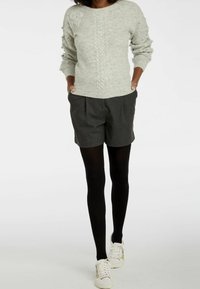 Hellgrauer Strickpullover mit strukturiertem Zopfmuster, kombiniert mit dunkelgrauen, maßgeschneiderten Shorts und schwarzen Strumpfhosen. Lässige weiße Sneaker runden den Look ab.