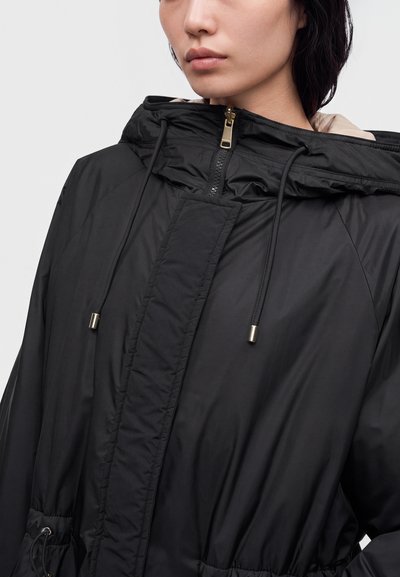 Femme portant une veste noire à capuche avec une fermeture éclair devant, des cordons et une taille froncée, vue de la poitrine vers le haut sur un fond uni.