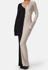 Femme portant une robe longue à côtes, bicolore, avec du noir à gauche et du beige à droite, boutonnée au centre avec de longues manches.