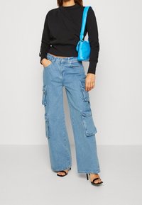 Ljust blå denim cargobyxor med vida ben, flera fickor och hög midja, parat med en figurnära svart långärmad topp och en blå handväska.