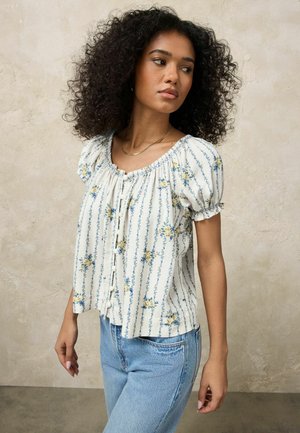 Frau mit lockigem Haar, die eine weiße Bluse mit Puffärmeln und Blumendekor sowie hellblaue Jeans trägt, steht vor einer beigen, strukturierten Wand.