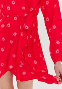 Červené květinové zavinovací šaty s dlouhými rukávy, detailem vázání v pase a volantem na lemu. Textilie má lehkou, hladkou strukturu s vzory sedmikrásek.