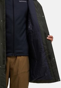 Persoană îmbrăcată cu pantaloni maronii și o jachetă bleumarin cu fermoar, ținând deschis un palton verde închis care arată căptușeala bleumarin matlasată.