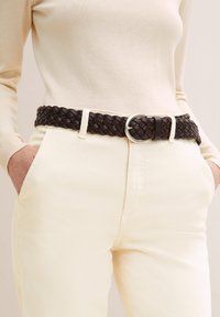 Cinturón de cuero trenzado marrón con hebilla circular plateada, usado sobre pantalones color beige claro. El cinturón presenta un diseño texturizado y trenzado.