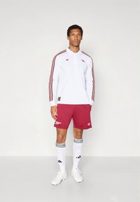 Camisola branca de manga comprida com detalhes vermelhos e três riscas vermelhas; calções bordô; meias brancas com riscas pretas; chuteiras de futebol brancas.