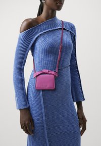 Donna che indossa un vestito blu a costine con spalle scoperte e una piccola borsa a tracolla viola con un dettaglio a fiocco sul davanti.