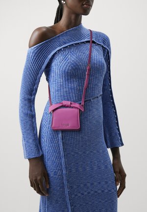 Kvinde iført en blå ribbet off-shoulder kjole og en lille lilla crossbody taske med en sløjfedetail foran.
